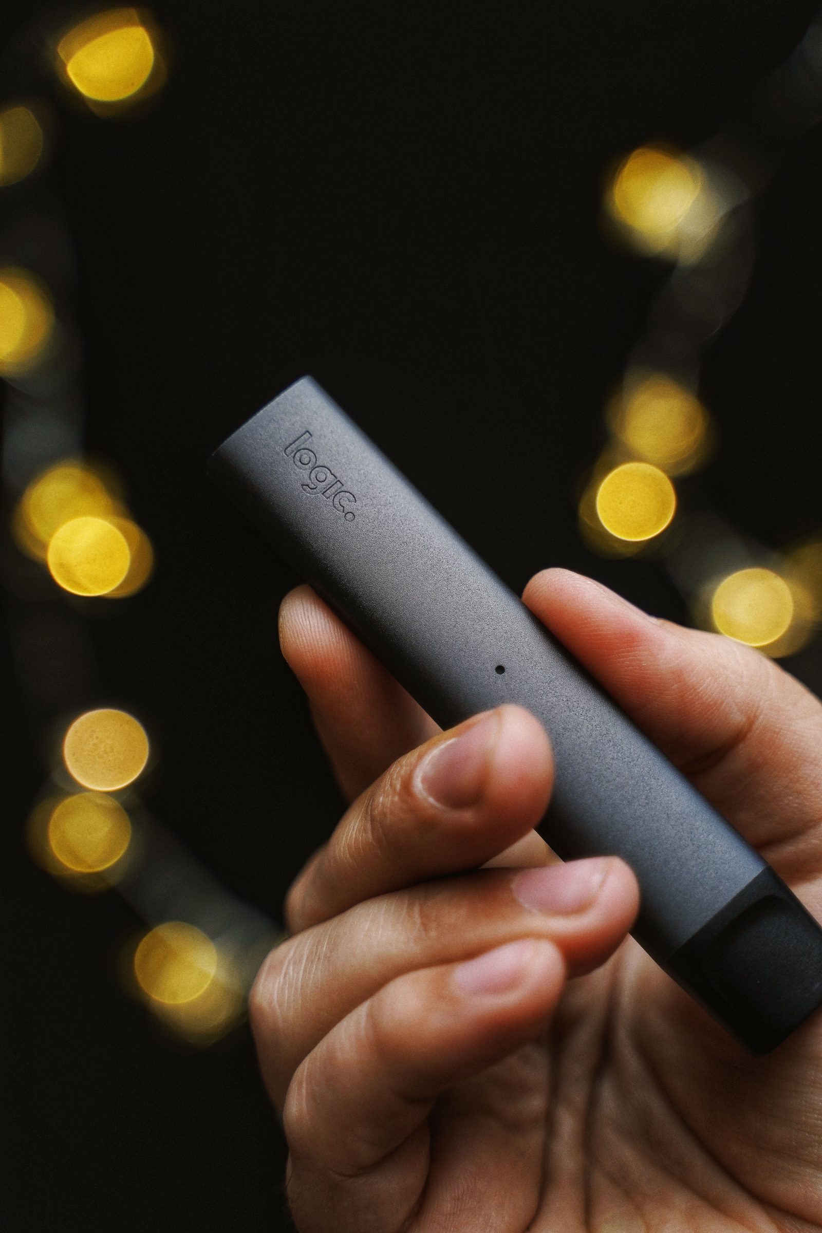 A hand holding a slim disposable vape device against warm background lights.